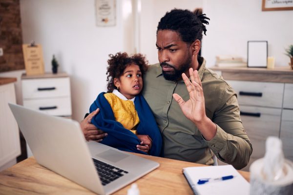 Little black girl wrapped in blanket sitting on father's lap while he is having video call over laptop at home.