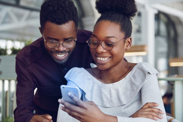 Happy African female in spectacles types message on modern smart phone while her boyfriend stands near her and looks at screen, have broad smile and white teeth. Dark young skinned woman and man