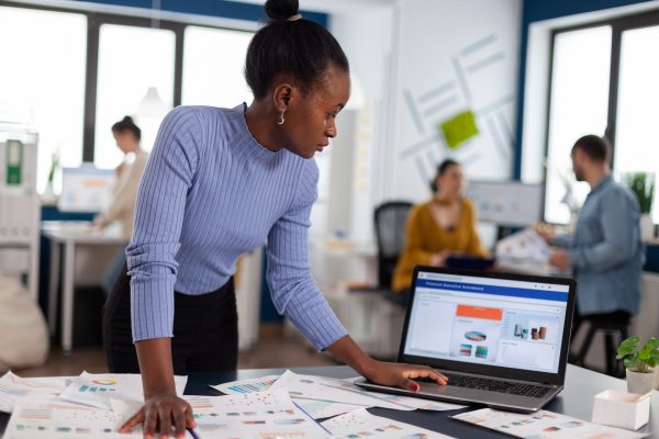 African woman scorlling on laptop looking concentrated at charts and multiethnic colleagues working on marketing. Diverse team of business people analyzing company financial reports from computer.