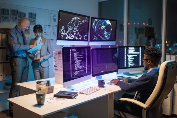 Young African businessman looking at data on computer screen while sitting in armchair in front of several monitors and typing